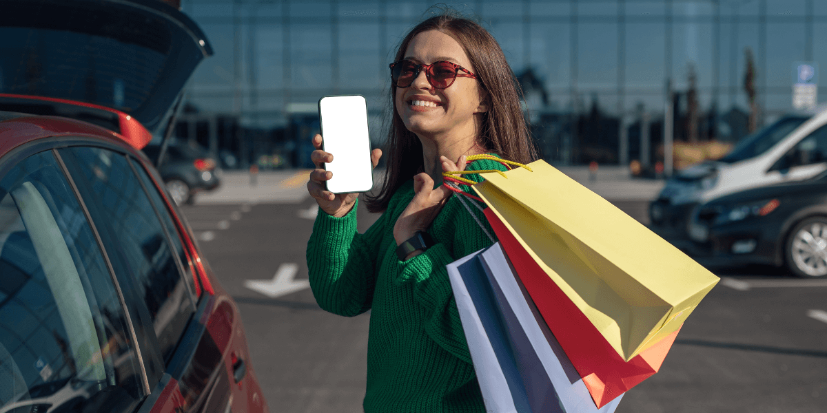 A young lady smiling and feeling happy while holding shopping bags beside the car, enjoying a successful shopping trip.